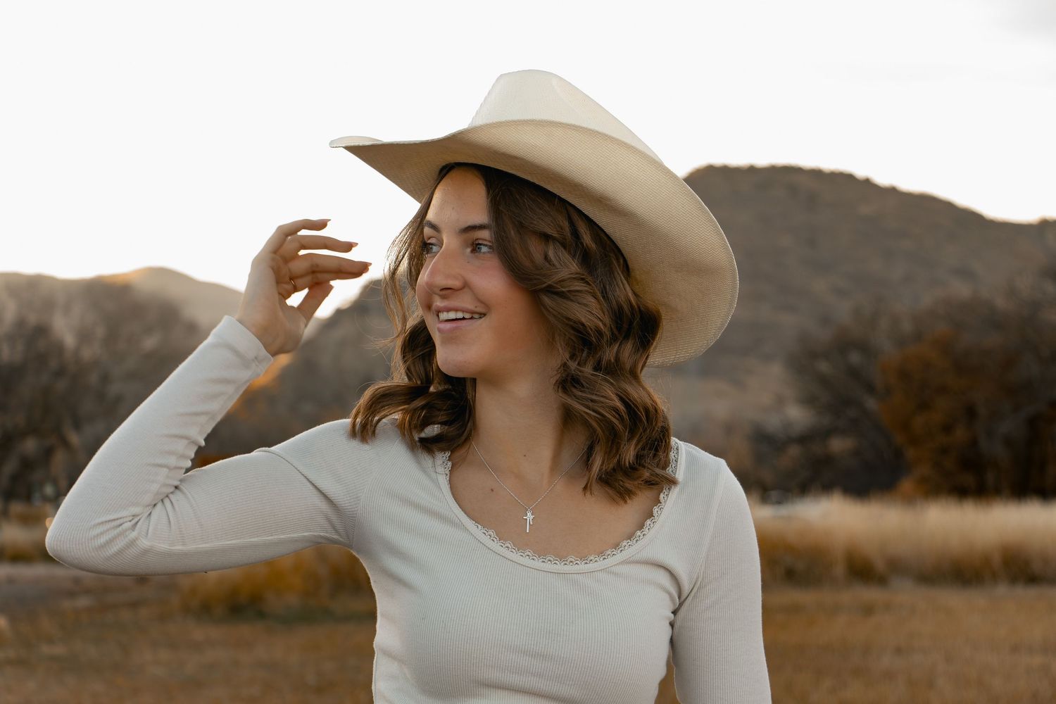 A person in a white dress and cowboy hat poses against a desert landscape during golden hour.