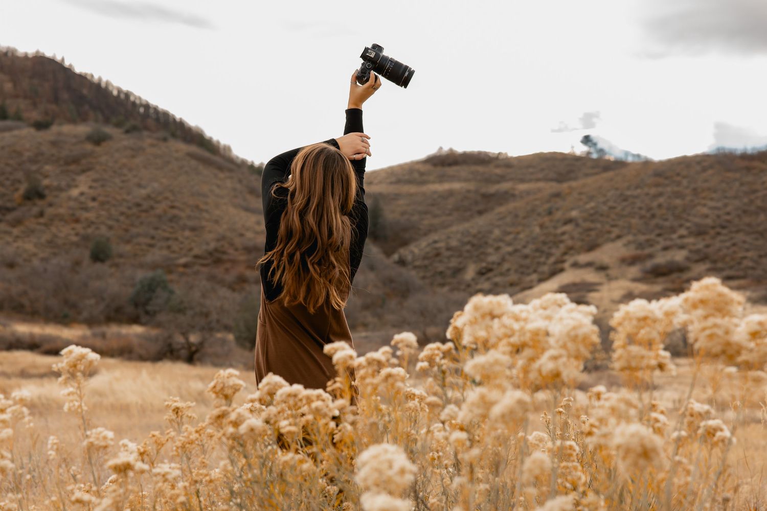 Woman holding camera above her head in golden field with dry wildflowers and rolling hills in background.
