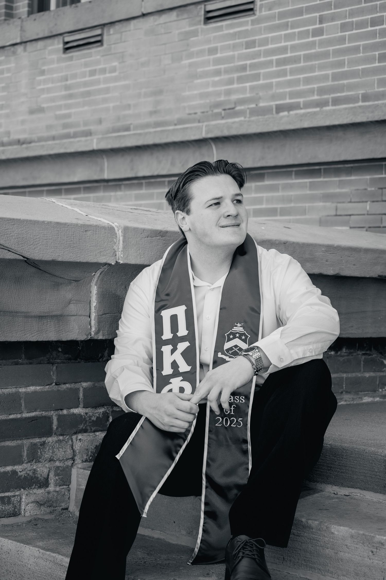 A graduate in cap and gown sits on brick steps while holding a guitar in black and white photography.