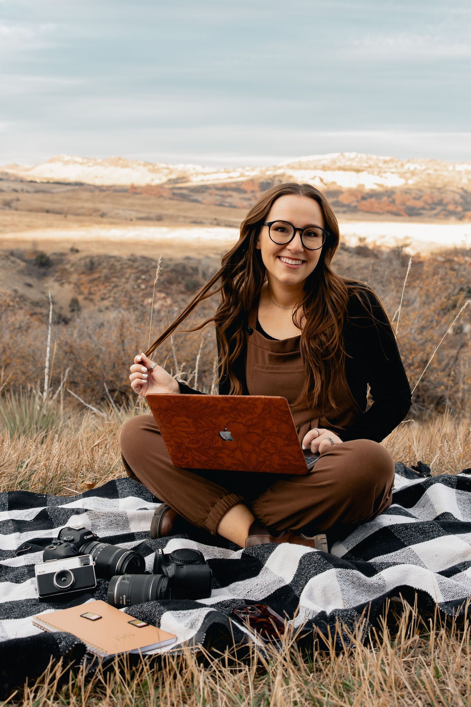 Remote worker with laptop sits on blanket in scenic mountain landscape during golden hour sunset.