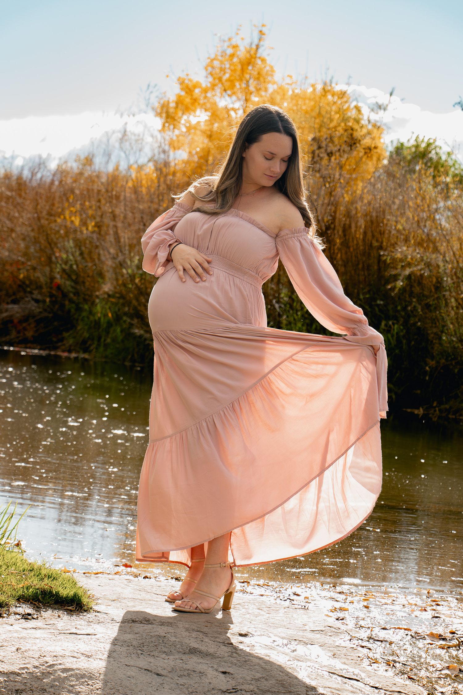 A woman in a flowing peach dress twirls gracefully by a lake during golden hour, creating an ethereal autumn scene.