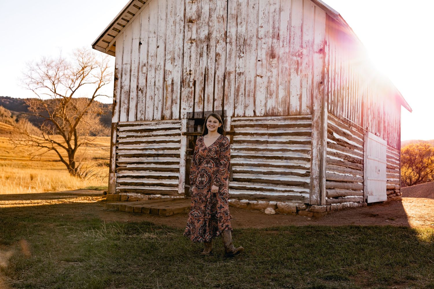 Woman in floral maxi dress poses by rustic white barn at golden hour on a rural farm.