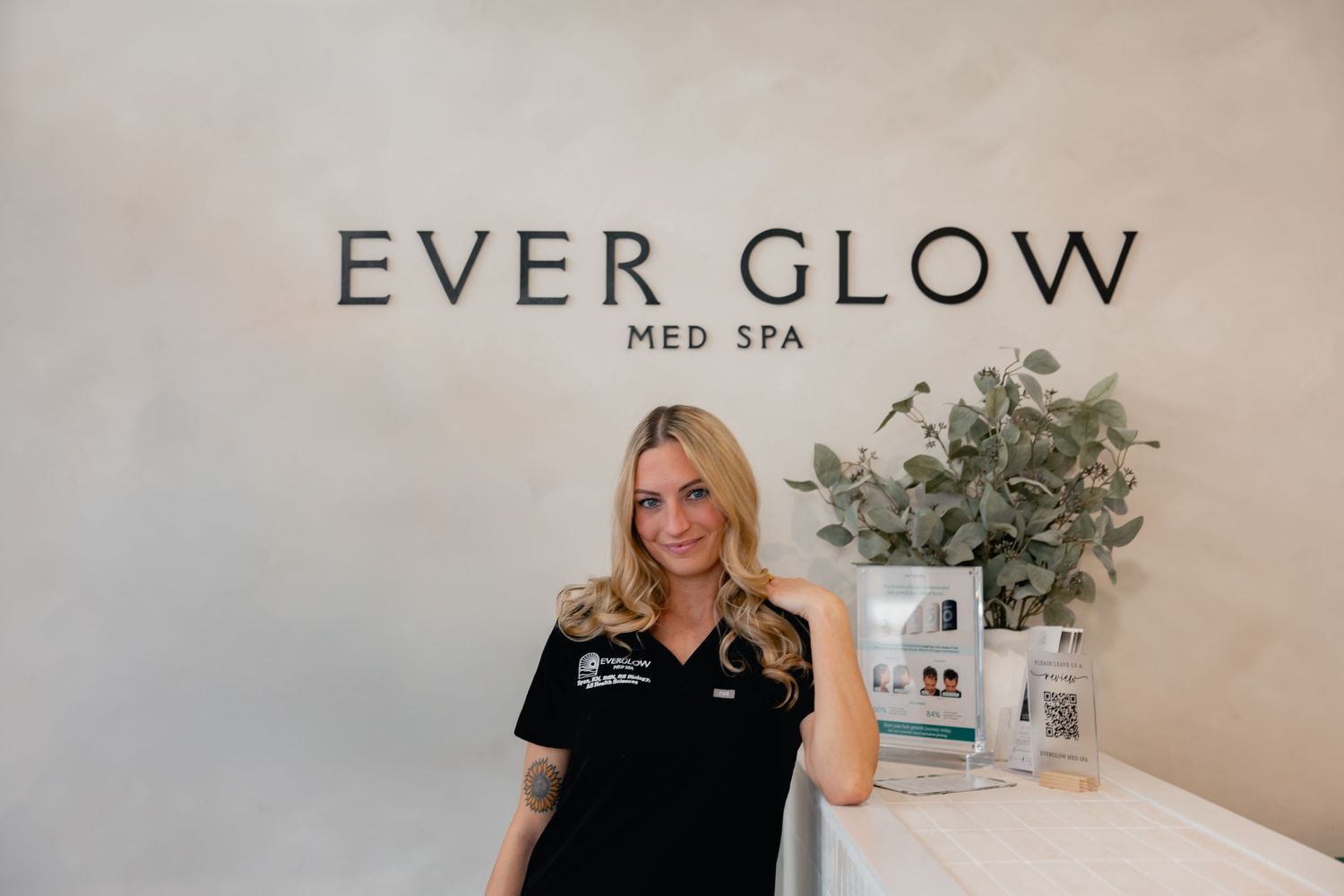 A smiling woman in black scrubs stands at the reception desk of Ever Glow Med Spa.