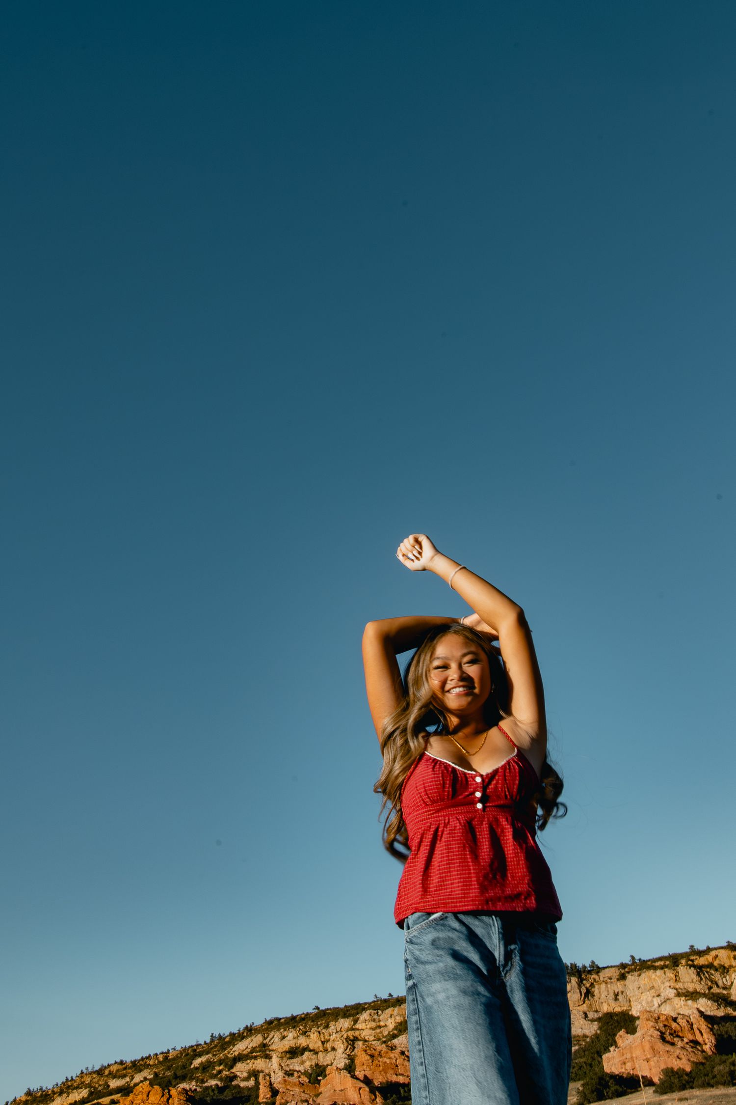 A person in a red tank top and jeans stretches their arms up against a vivid blue sky and rocky landscape.