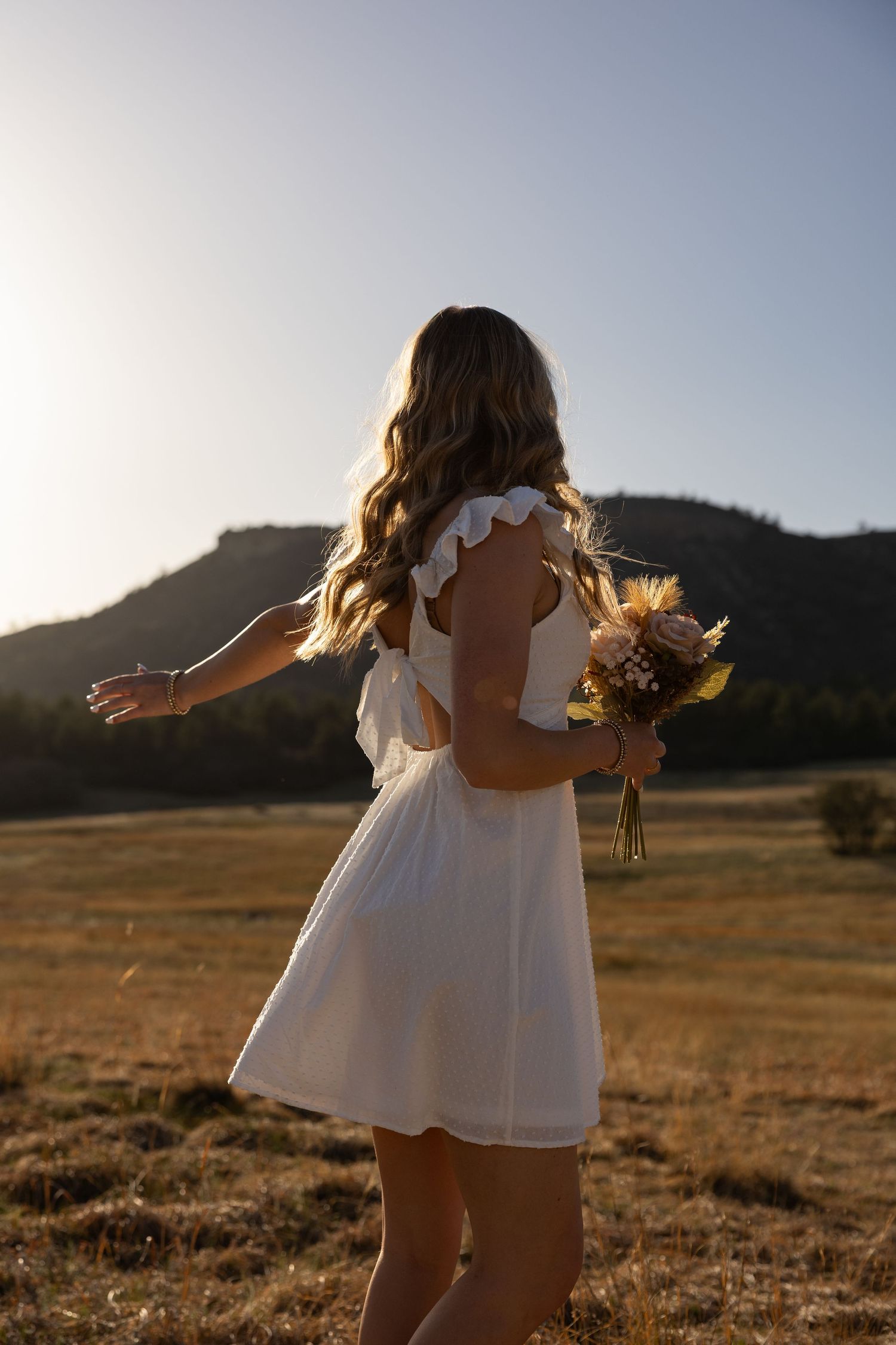 Woman in white dress holding bouquet in golden field at sunset with mountain backdrop.