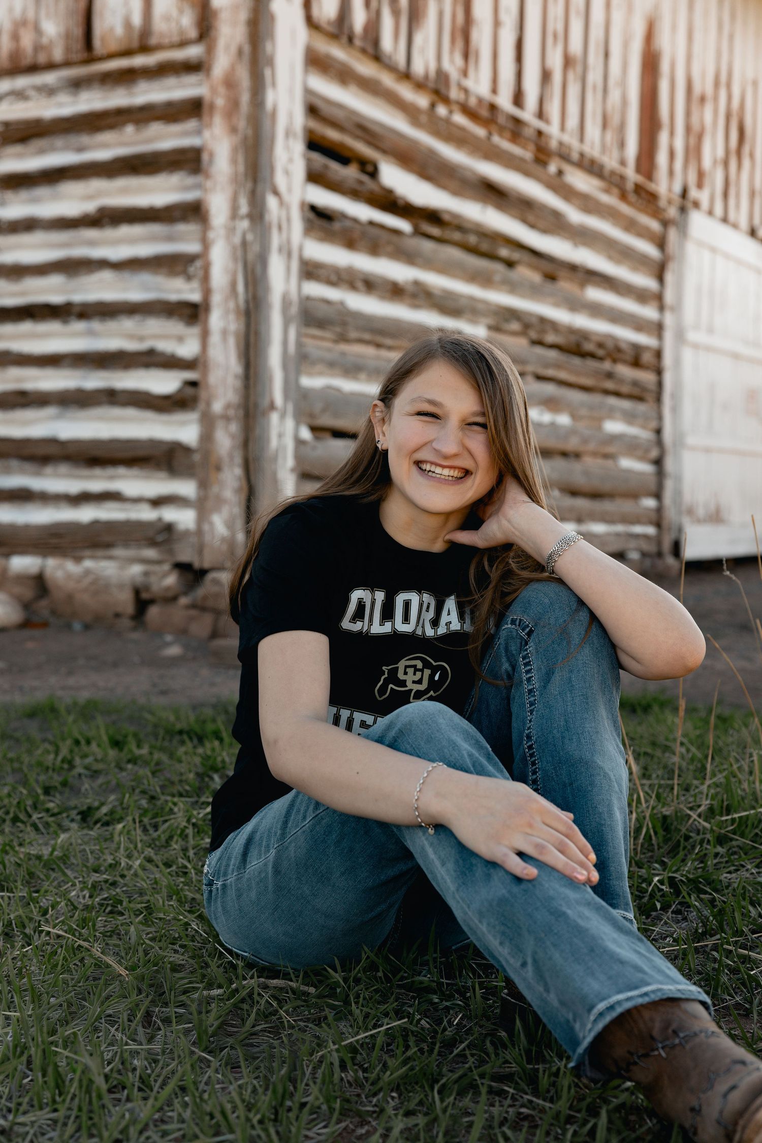 Smiling young woman in Colorado Buffaloes shirt and jeans sitting on grass beside rustic wooden barn.