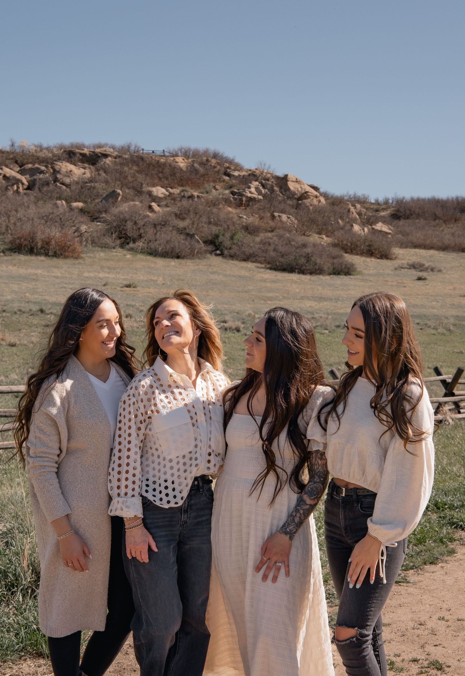 Four friends in casual outfits laughing together on a dirt path with a rocky hillside and blue sky in the background.