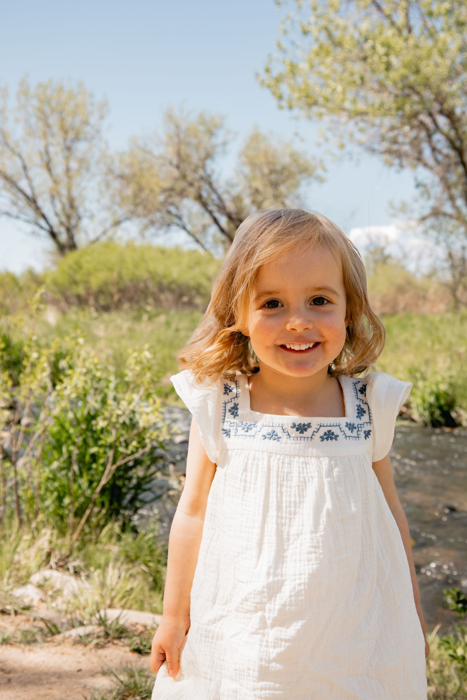 A young child wearing a white summer dress smiles brightly while standing outdoors in a natural setting with trees.