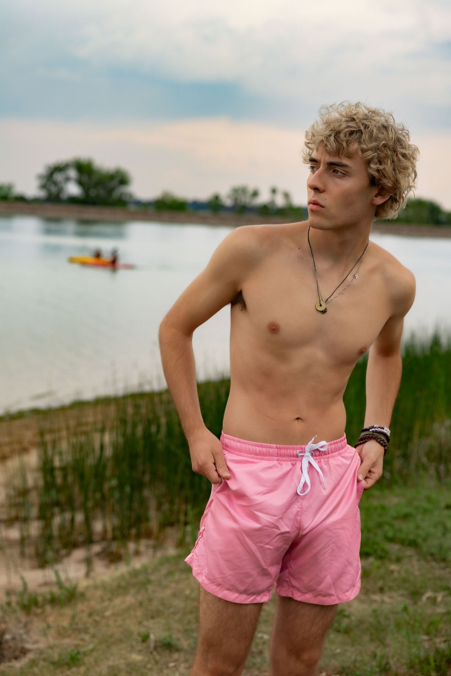 Young man with curly blonde hair in pink swim shorts standing by a lake with a kayaker in the background.