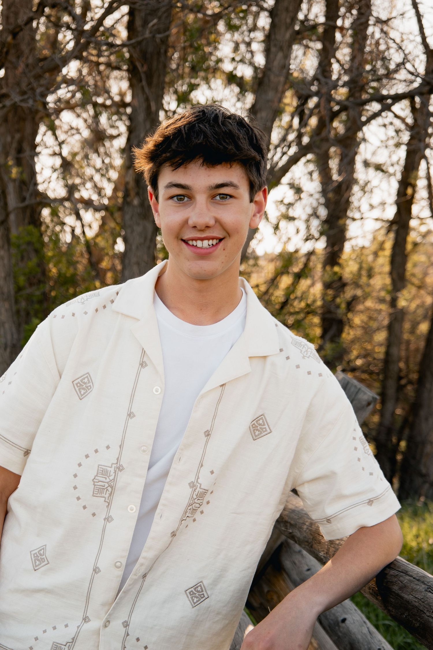Portrait in outdoor setting showing someone wearing a white patterned button-up shirt and t-shirt against wooded background.