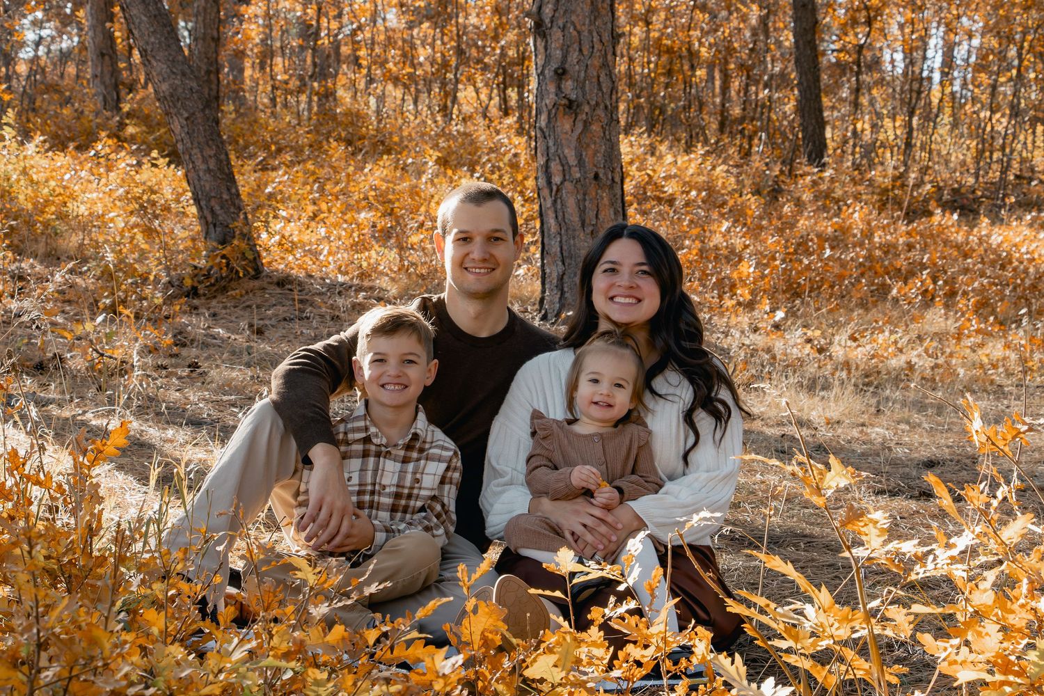A family sits together outdoors surrounded by golden autumn foliage in a wooded setting.