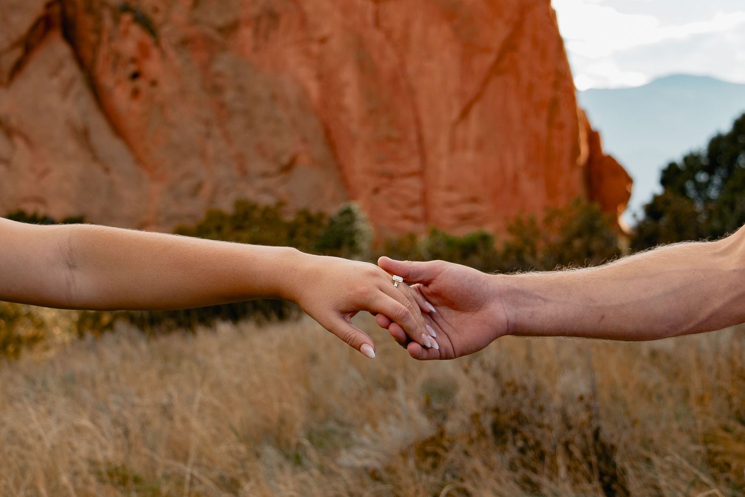 Two hands reach out to hold each other against a backdrop of red rock cliffs in a desert landscape.