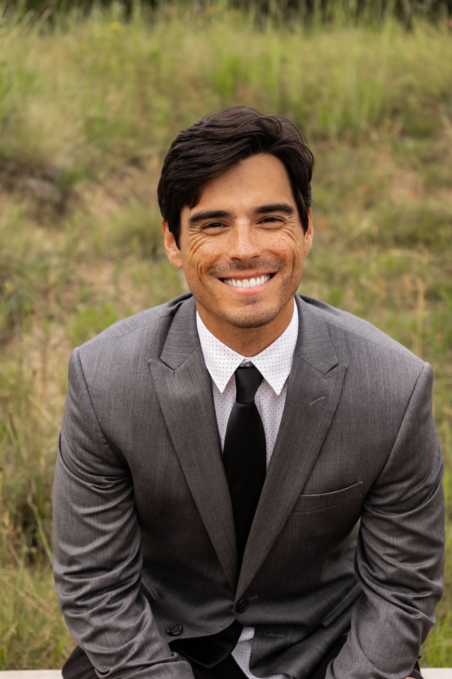 Headshot of professional in gray suit and black tie smiling warmly outdoors in natural setting.