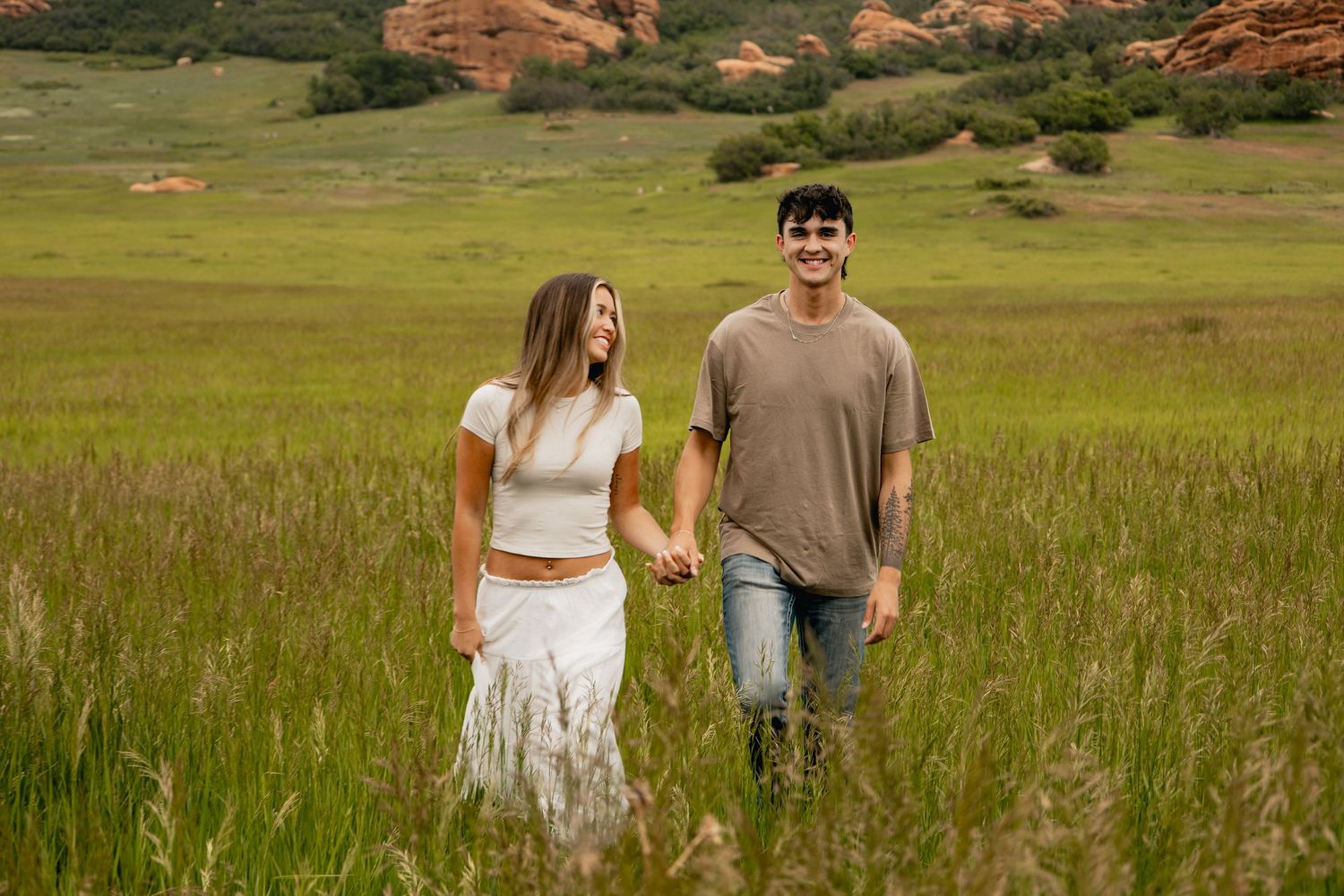 A couple holds hands while walking through tall grass in a scenic mountain meadow against red rock formations.