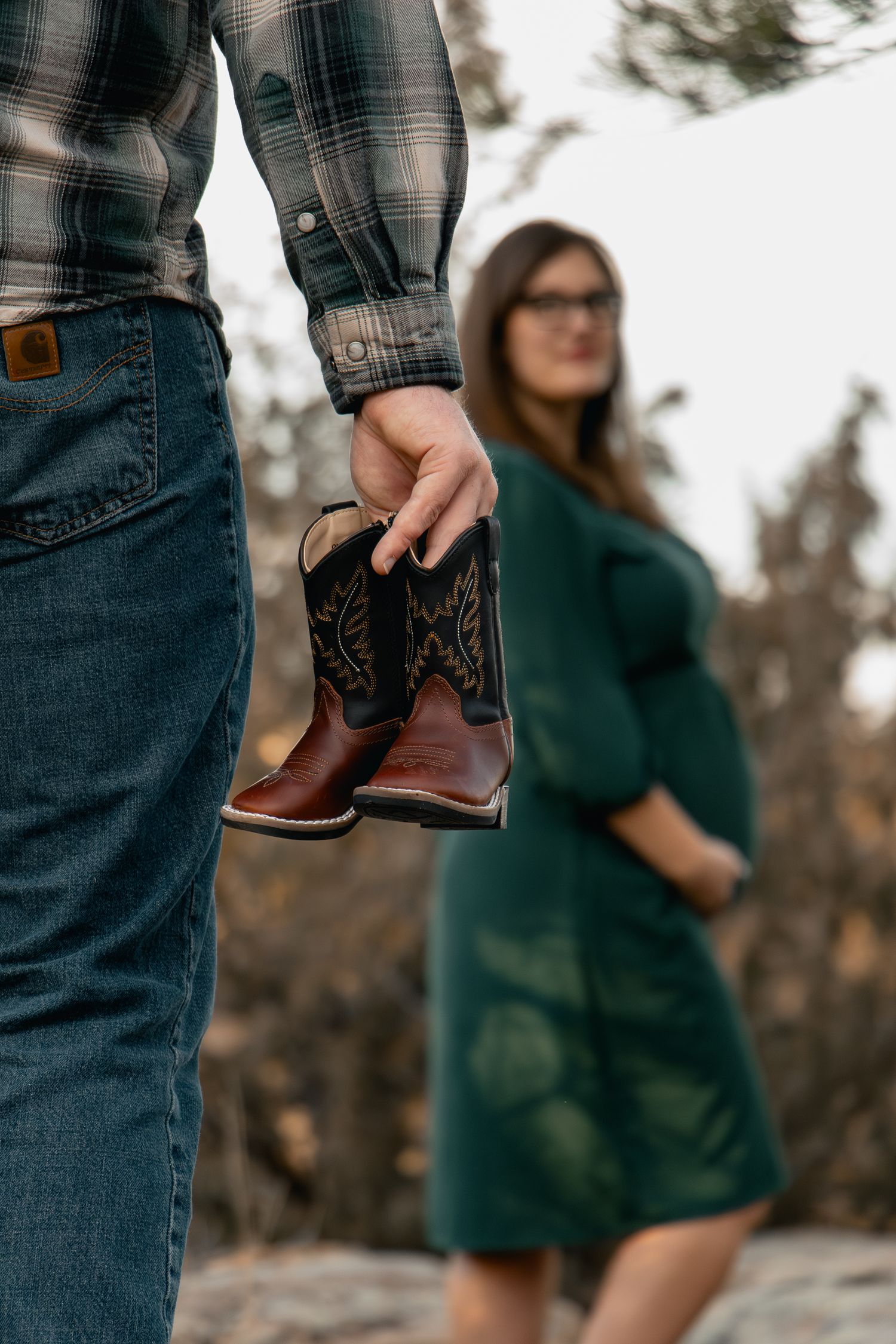 Man holding tiny cowboy boots with pregnant woman in green dress standing in background outdoors.