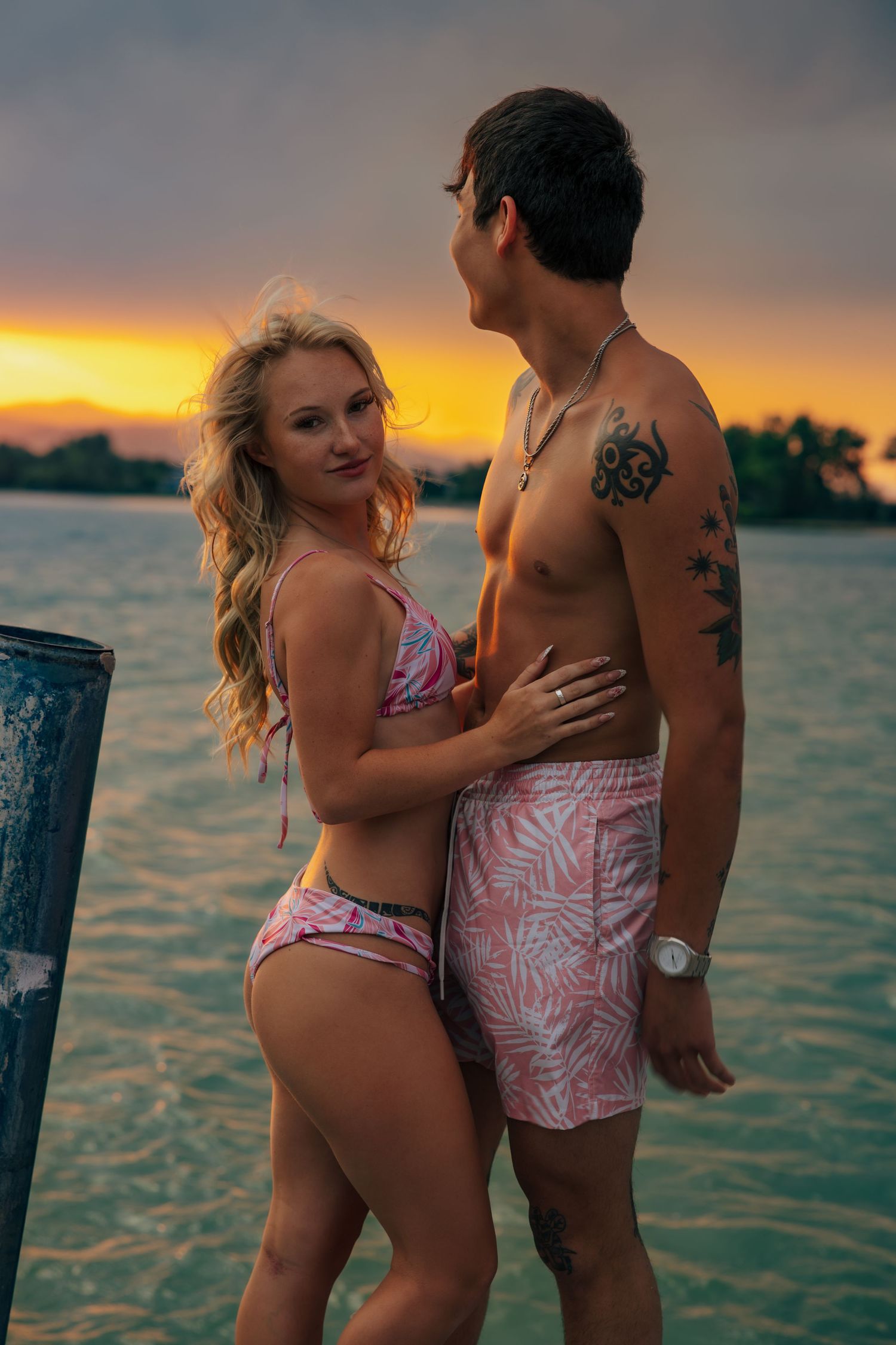 Couple in matching pink swimwear embracing on a dock at sunset over calm water.