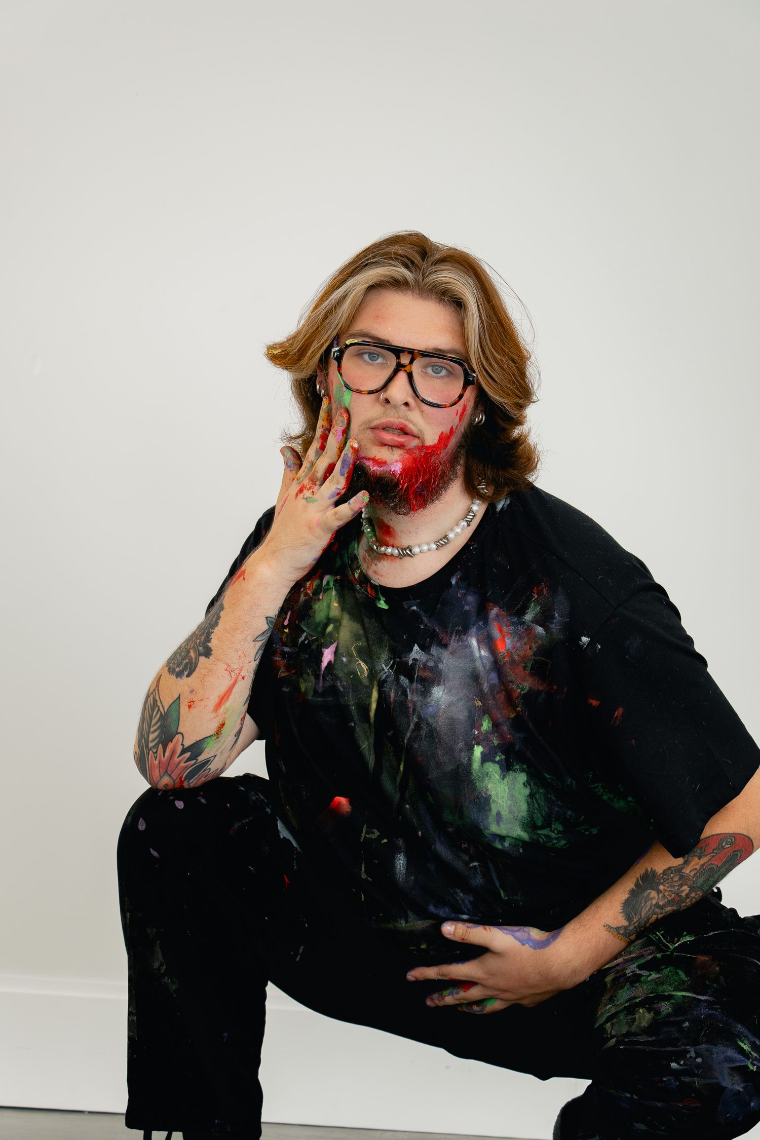Person in black clothing with tattoos poses playfully against white background with colorful paint marks.