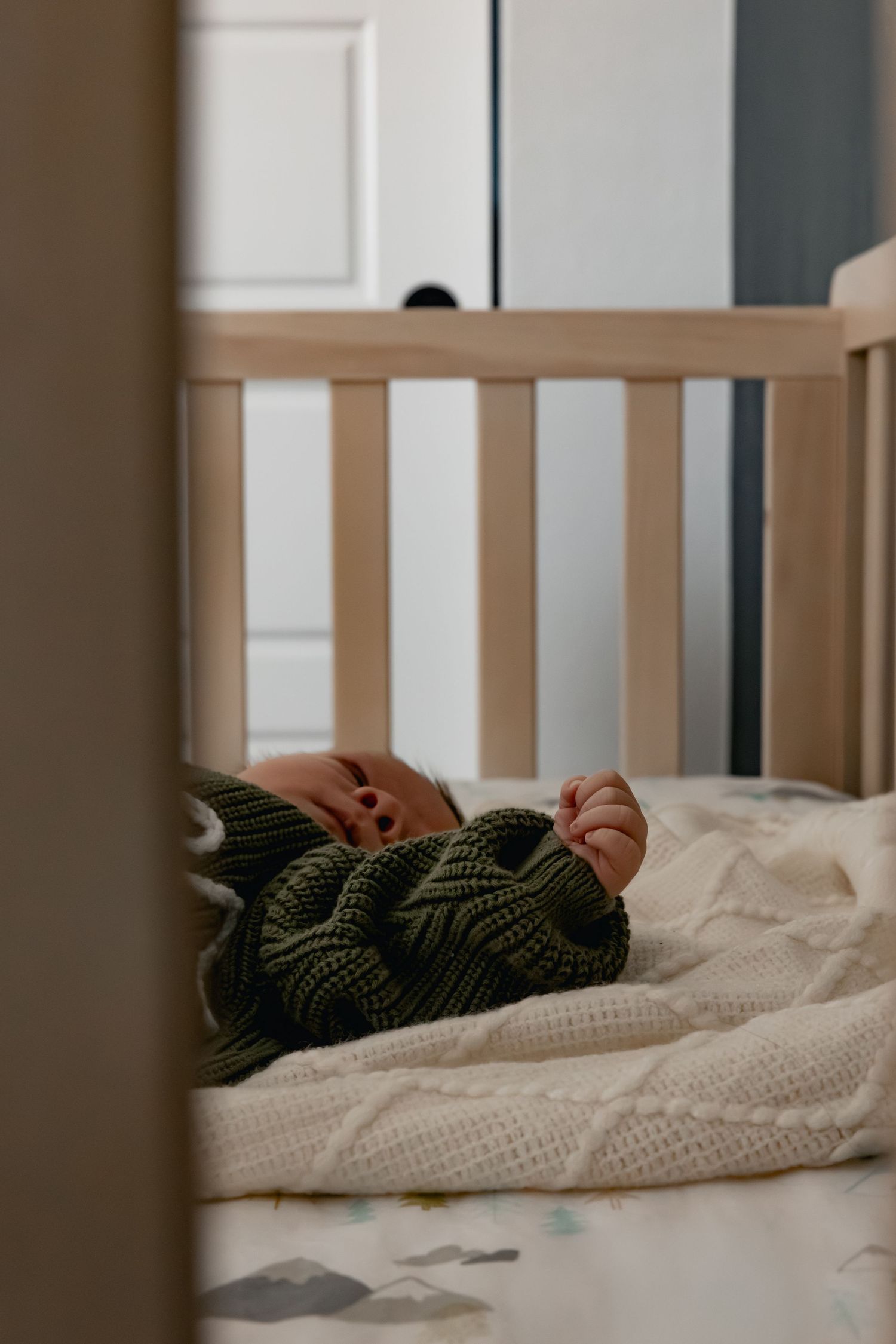 Baby sleeping peacefully in a wooden crib, wearing a green knit outfit on a white textured blanket.