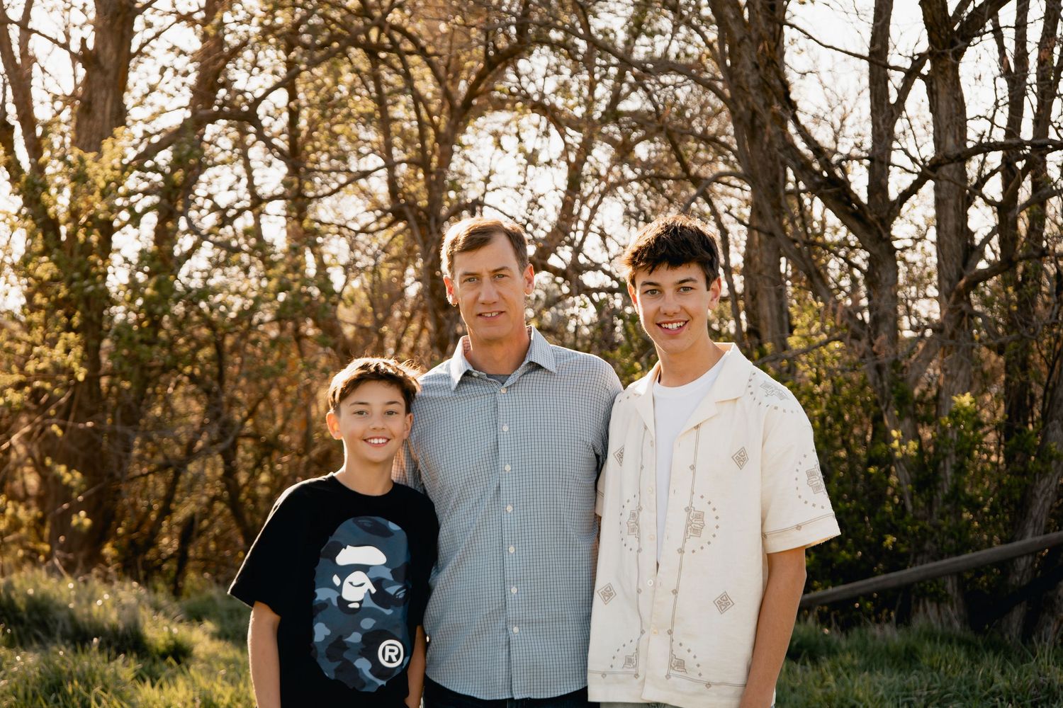 Three people stand together outdoors during golden hour with trees and natural light in the background.