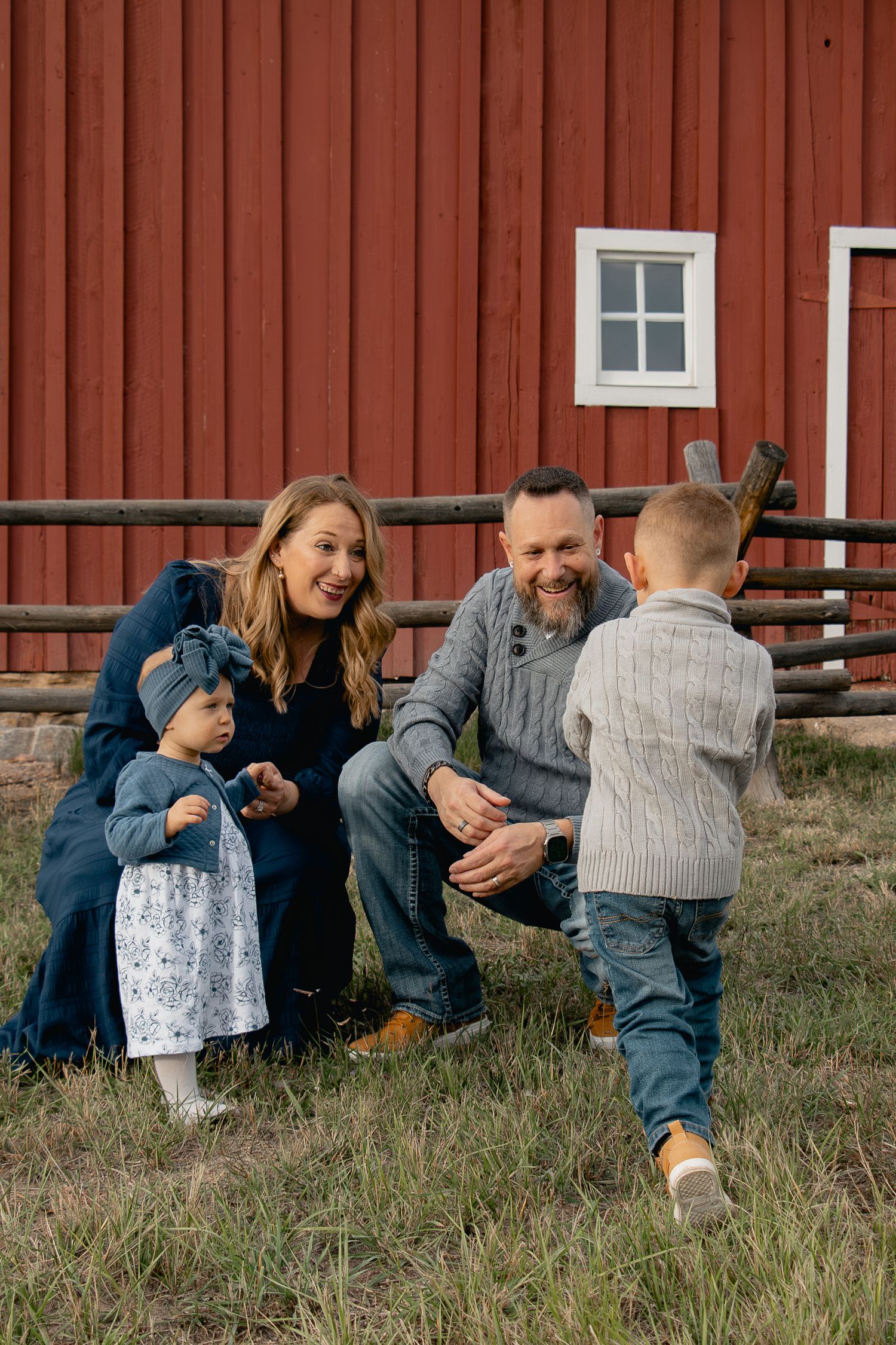Group of people sitting on grass against red barn wall having casual outdoor gathering.