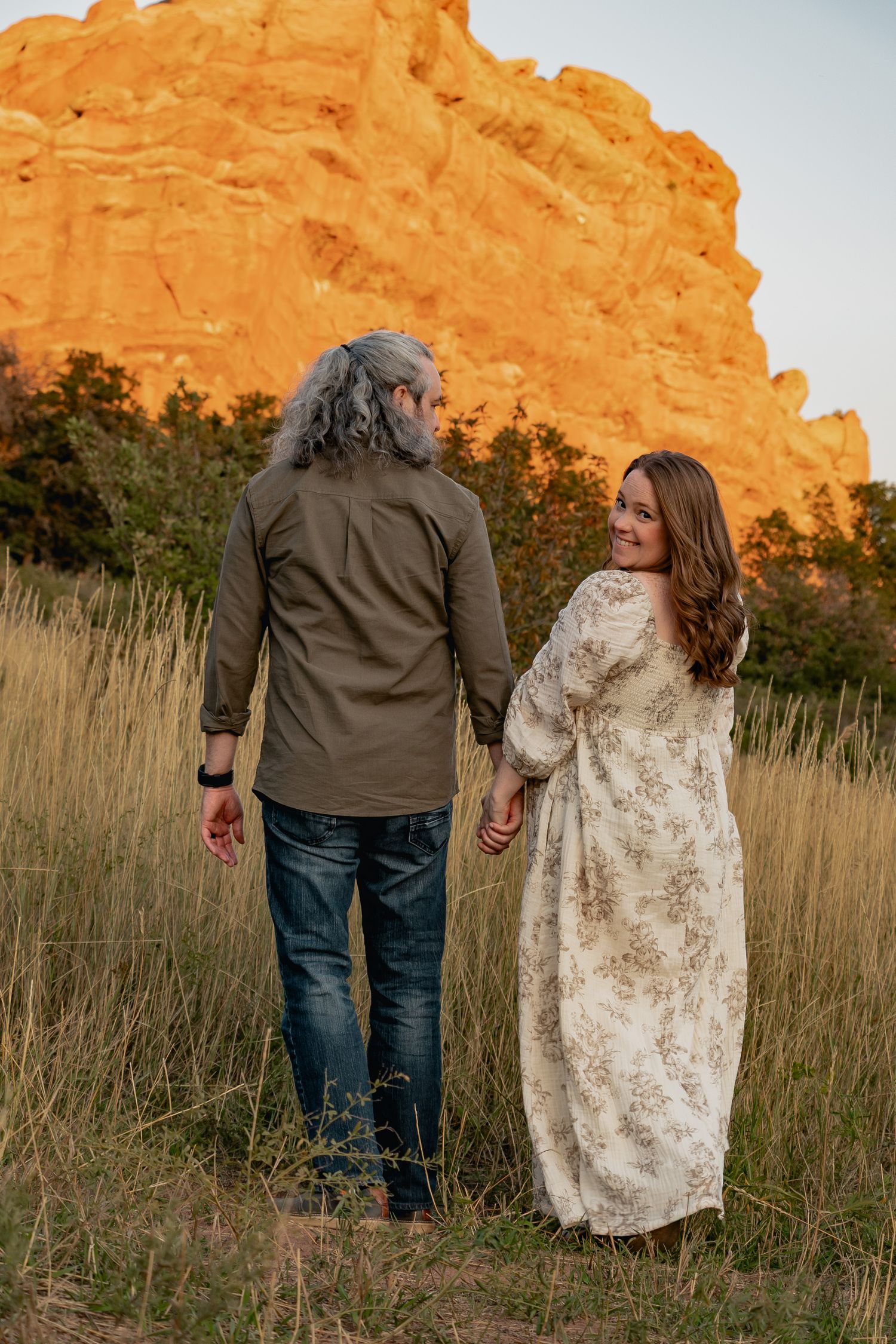 A couple holds hands while walking through tall grass with orange sandstone cliffs in the background during golden hour.