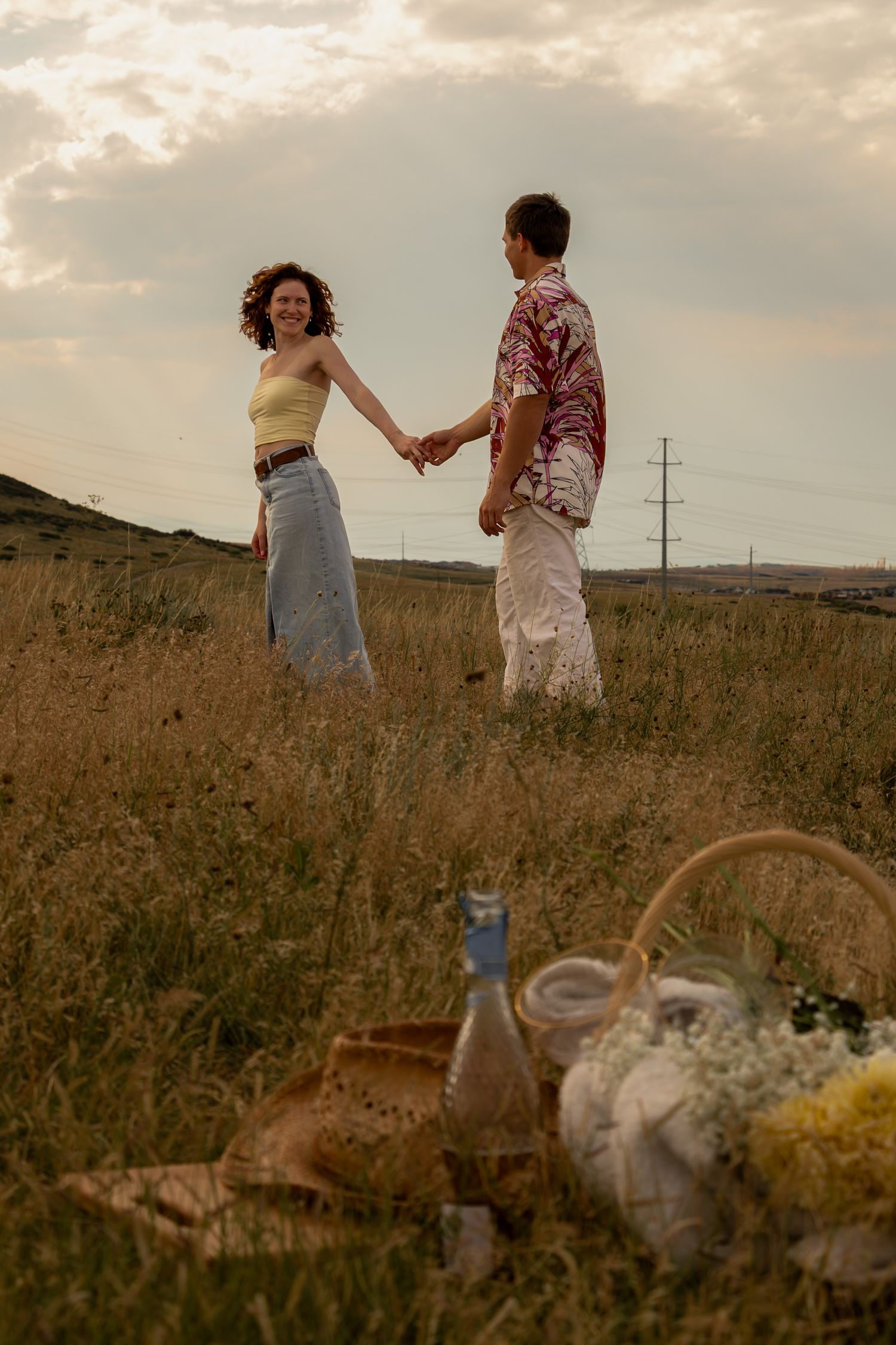 Two people in summer dresses stroll through a golden wildflower field at sunset with picnic items visible in foreground.