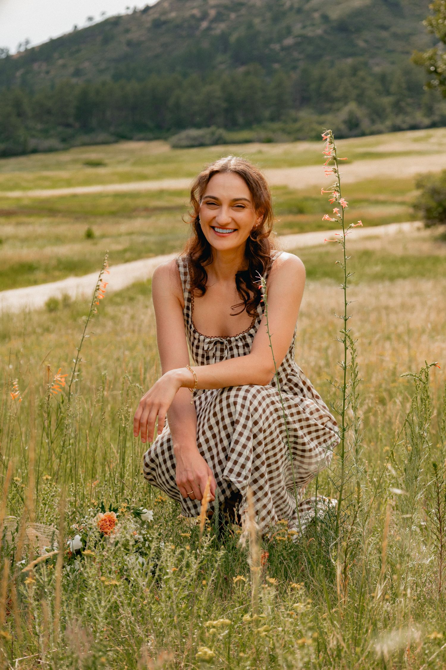 Someone wearing a gingham dress sits among tall grass and wildflowers in a mountain meadow on a summer day.