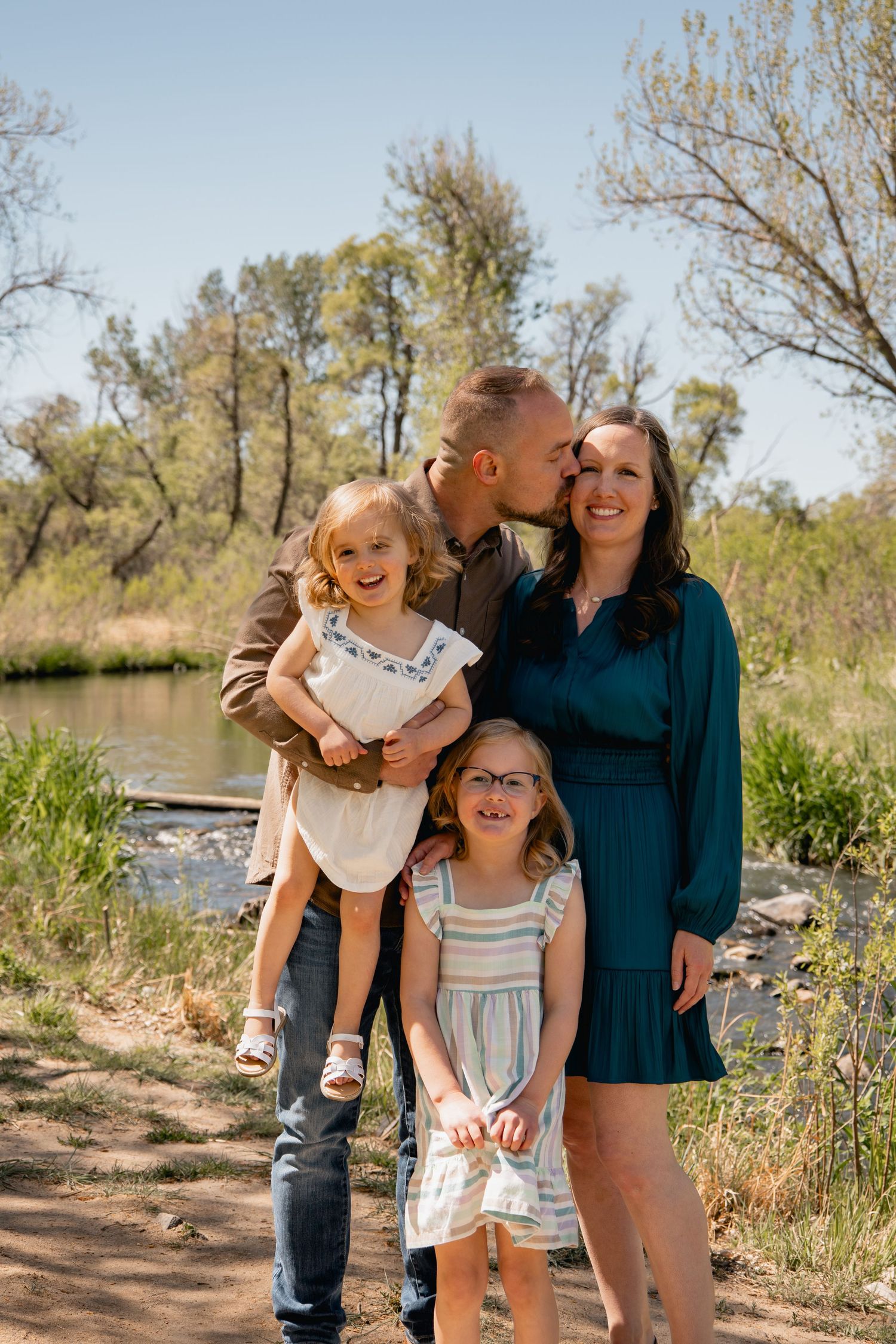 A family of four poses together on a sunny day by a river with trees in the background during a spring photoshoot.