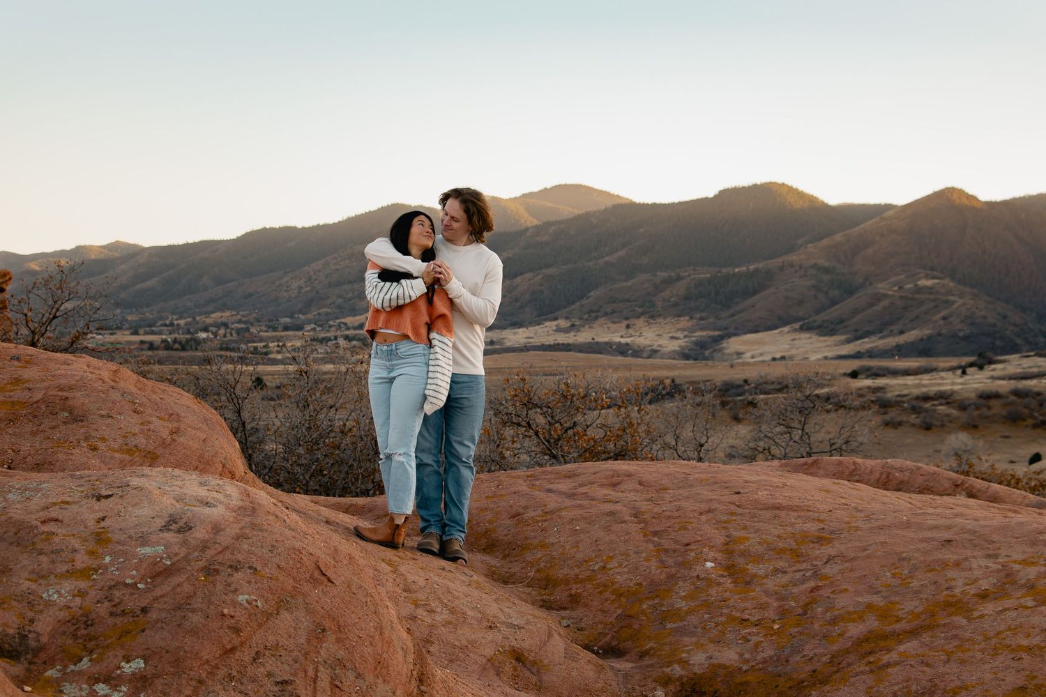 Couple embraces on red rock formations during golden hour in a desert landscape.