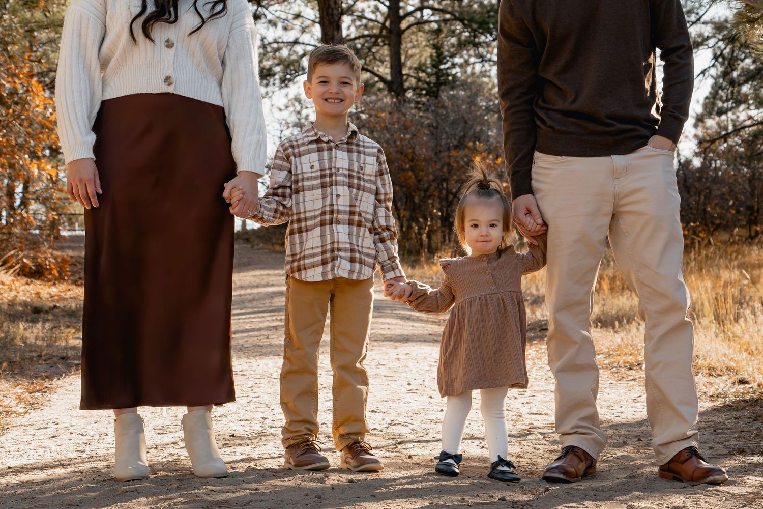 A family wearing coordinated earth-toned outfits stands together in warm autumn sunlight for outdoor portraits.