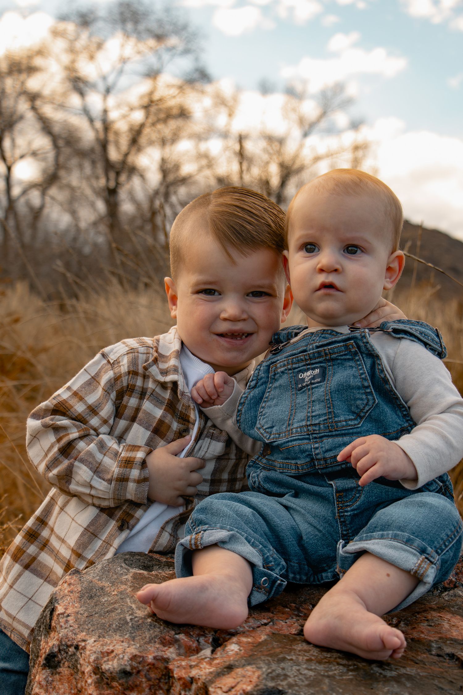 Two babies in plaid shirt and overalls sit together on a rock during an autumn outdoor photoshoot.