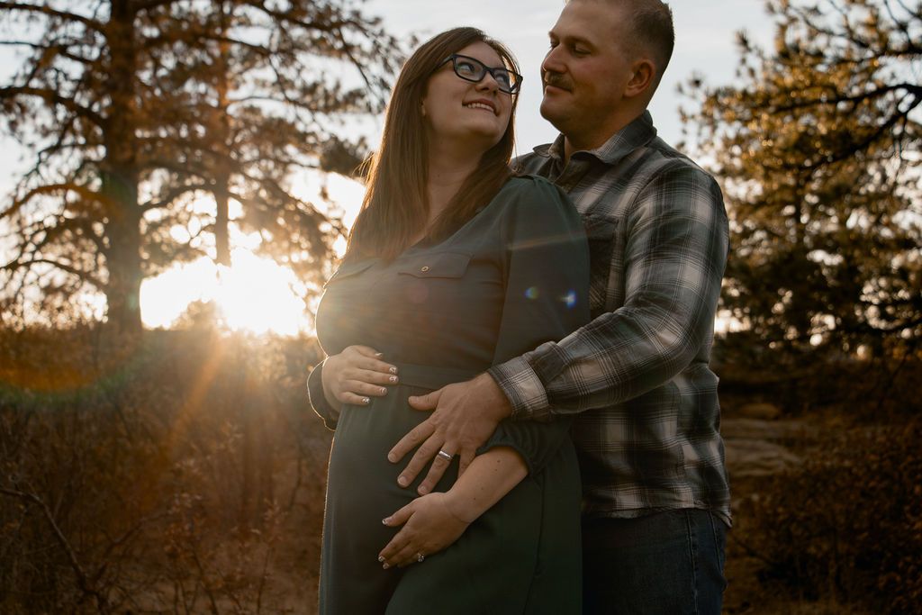 A couple embraces in a romantic sunset moment among tall evergreen trees during a golden hour photoshoot.