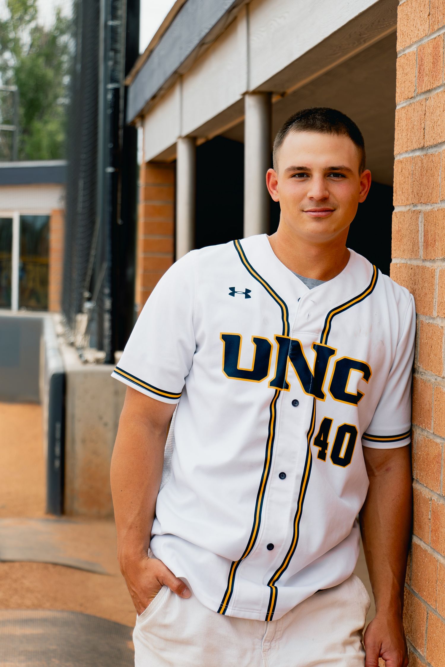 Baseball player in white UNC uniform number 40 stands against brick wall at athletic facility.