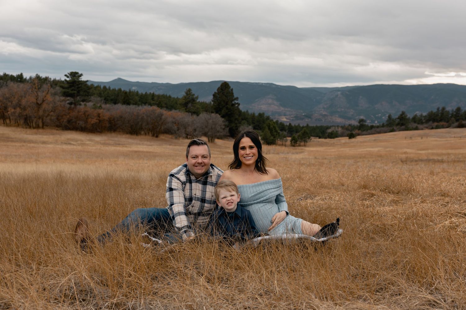 Family of three sitting in golden meadow with mountain backdrop on cloudy day.