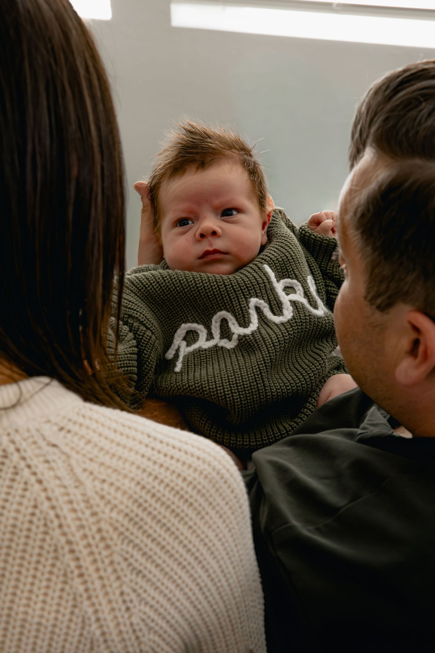 Newborn baby in green knit sweater held by parents, wearing a park-themed outfit in a cozy indoor setting.