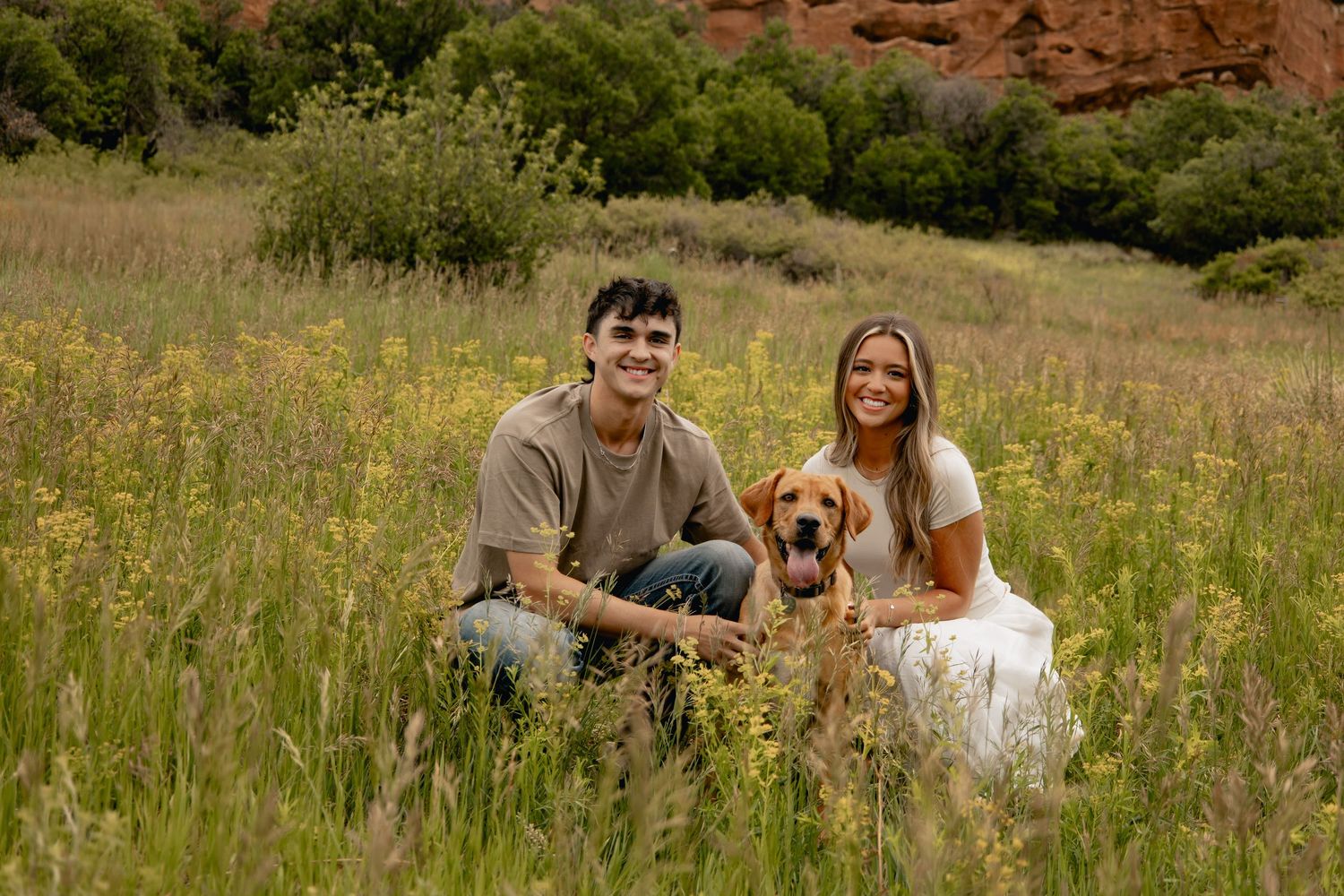 A couple sits with their golden dog in a sunny meadow with red rock formations in the background.
