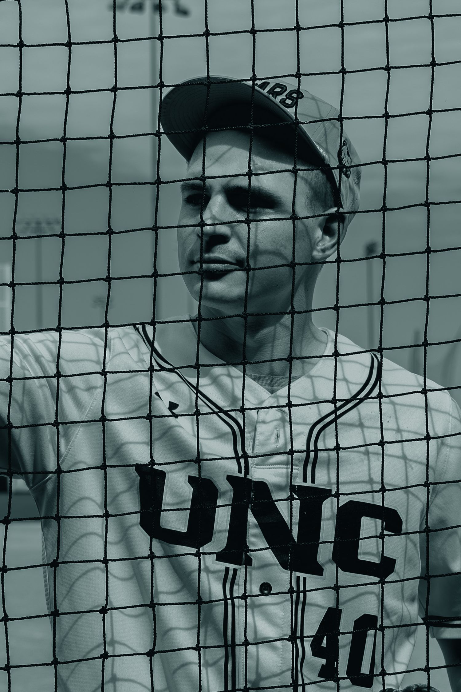 Black and white photo of a baseball player in a UNC uniform standing behind protective netting at a baseball field.
