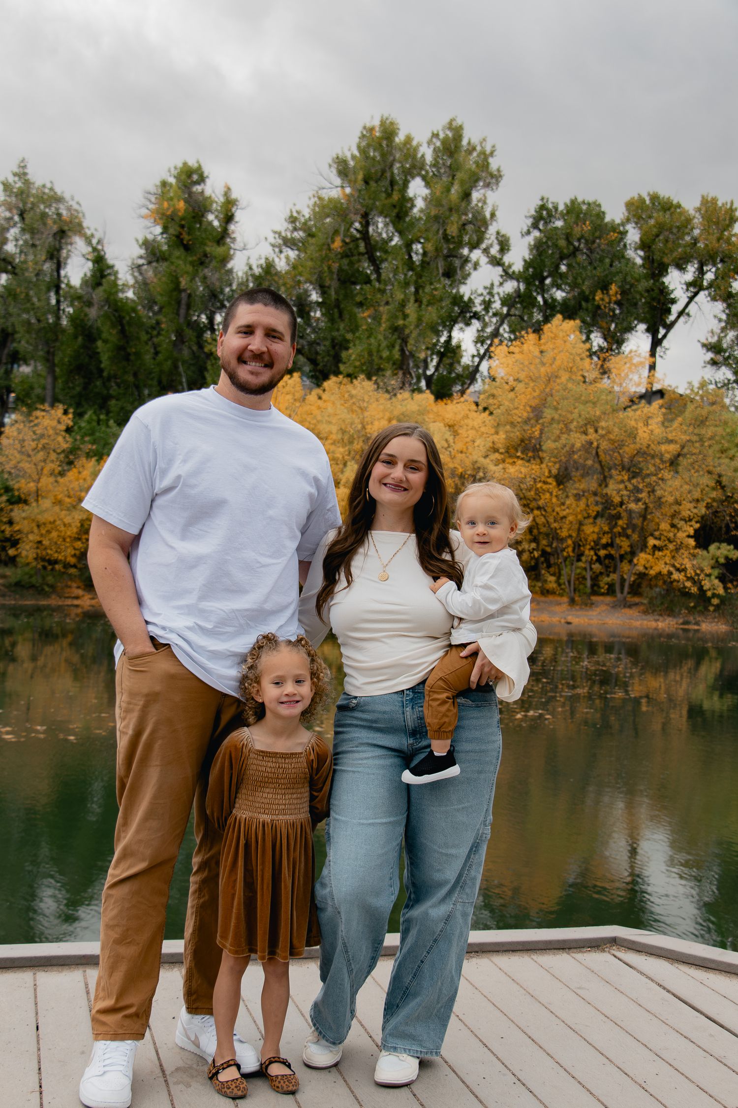 A family poses together on a wooden dock by a lake with autumn trees in the background.