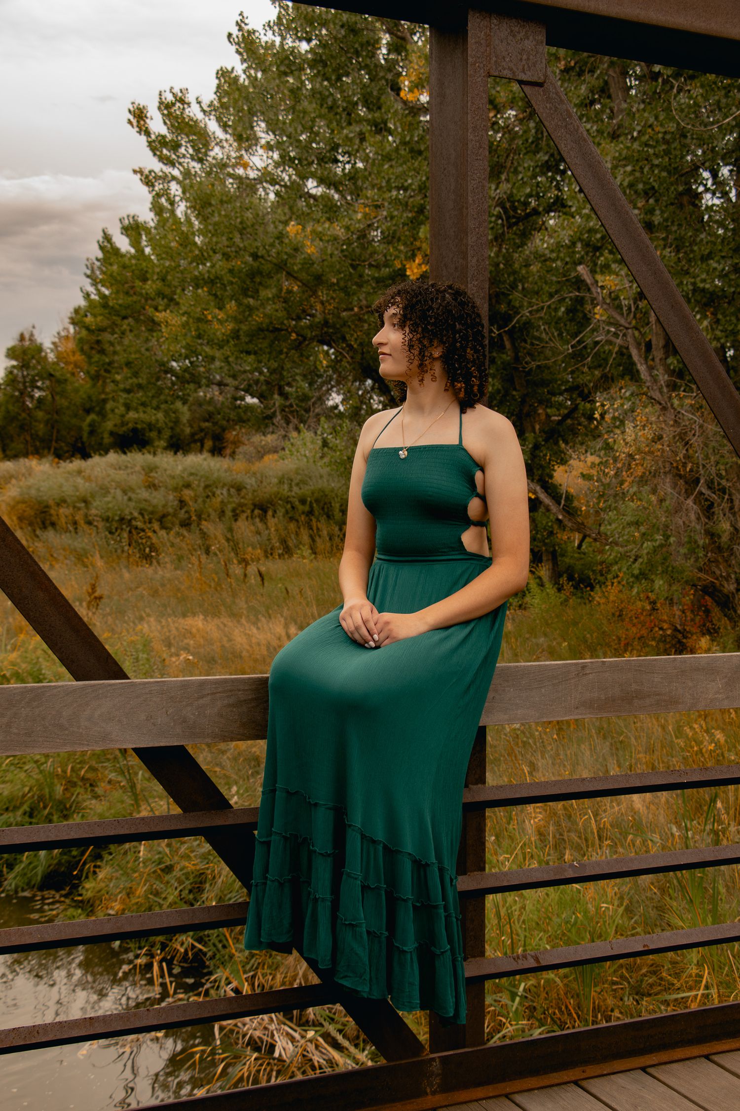 A person in an emerald green dress poses thoughtfully on a wooden bridge railing at sunset.