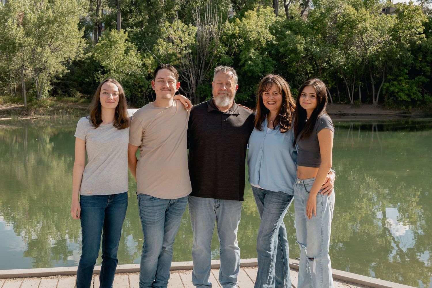 A group wearing casual jeans and tops poses together outdoors by a serene lake on a sunny day.
