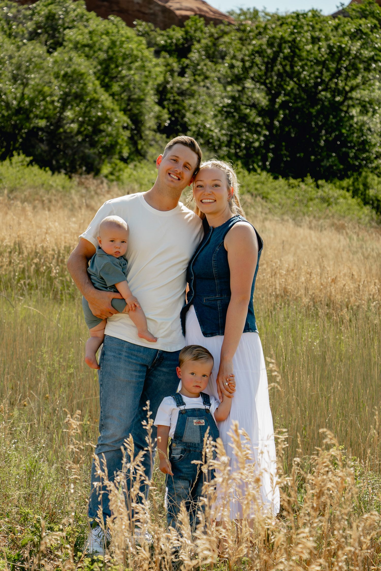 A smiling family poses together in a sun-lit meadow with tall grasses and trees in the background.