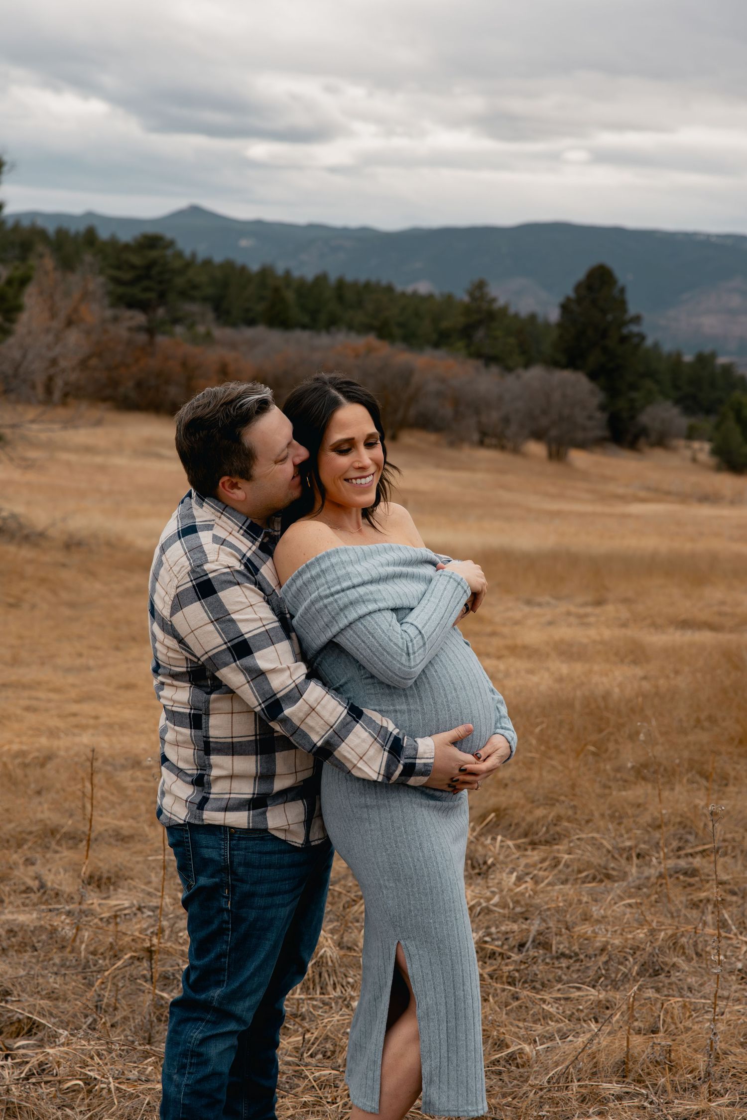 Couple embracing in golden field with mountain backdrop during maternity photo session, woman in blue off-shoulder dress.