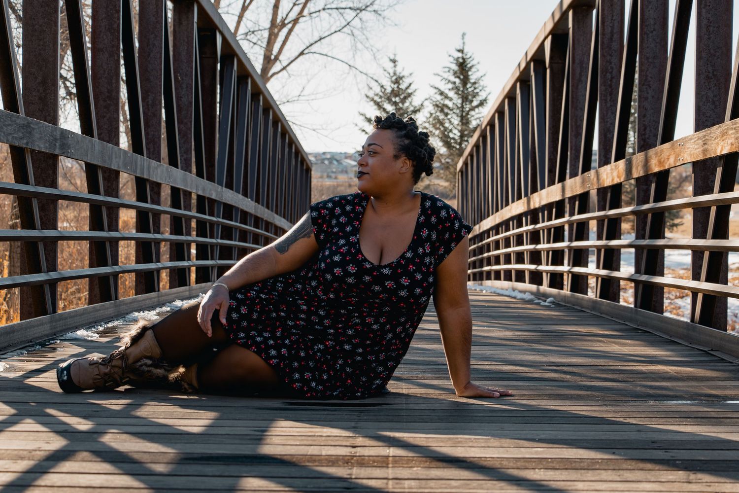 Woman in floral dress sitting on wooden bridge, posing outdoors on sunny day.