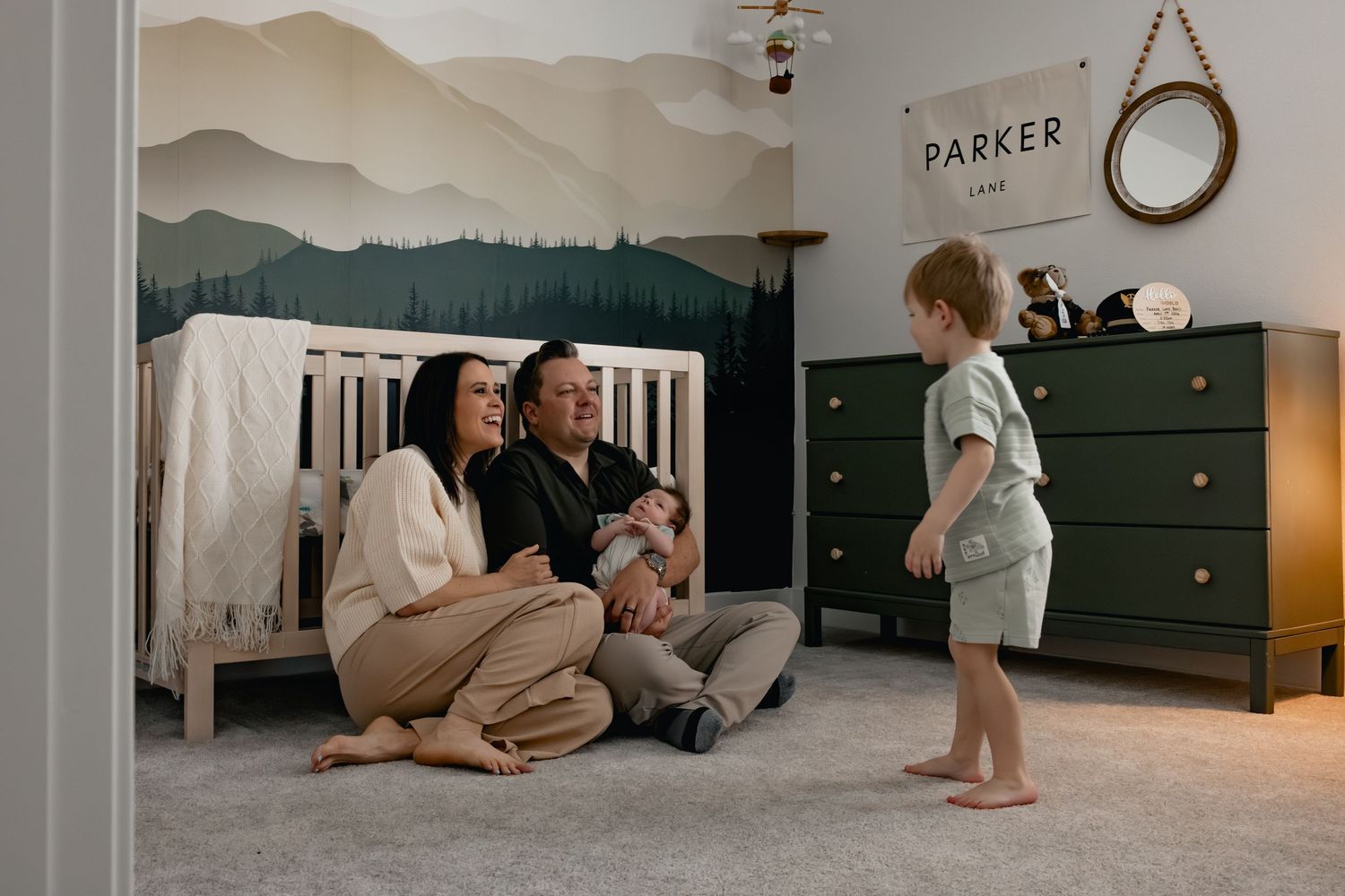 Parents sit on nursery floor with newborn while toddler stands nearby, mountain mural and crib in background.