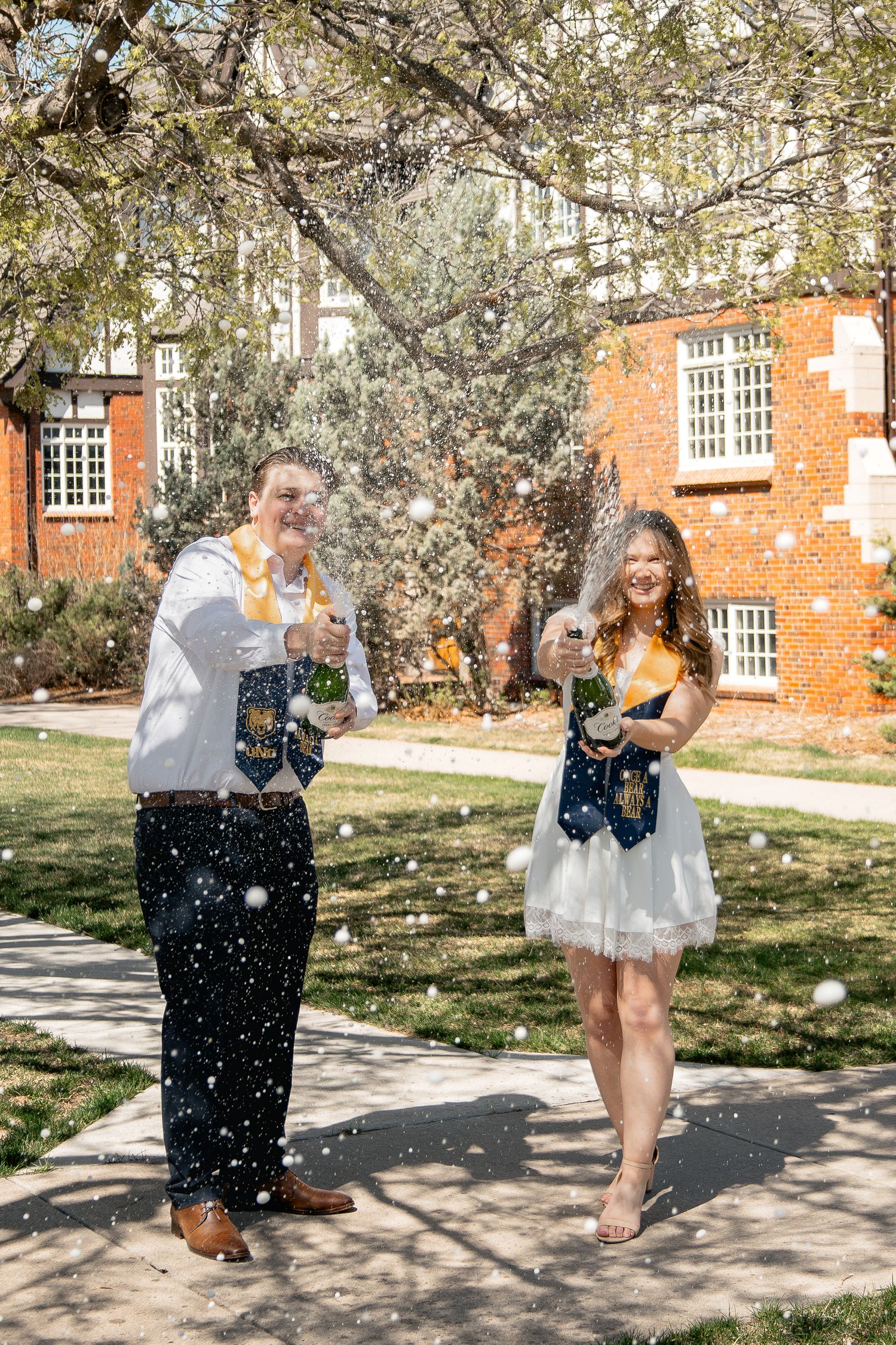 Two people in formal attire popping champagne bottles under blooming cherry blossoms near a brick building.