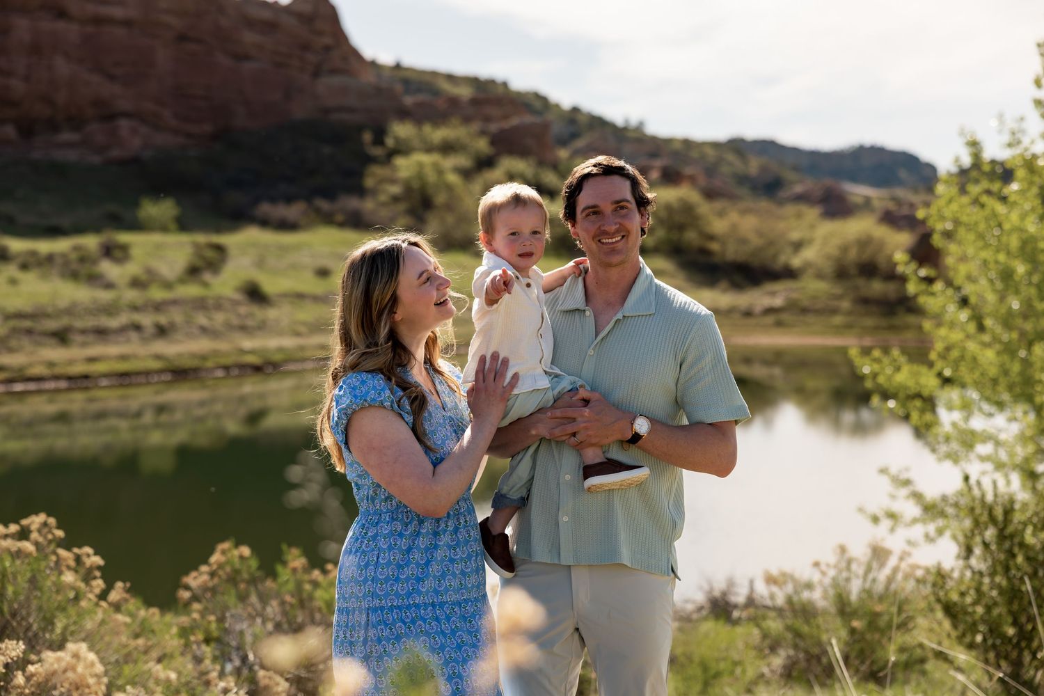 Happy family with baby posing outdoors near a scenic lake with red rock cliffs and green hills in background.