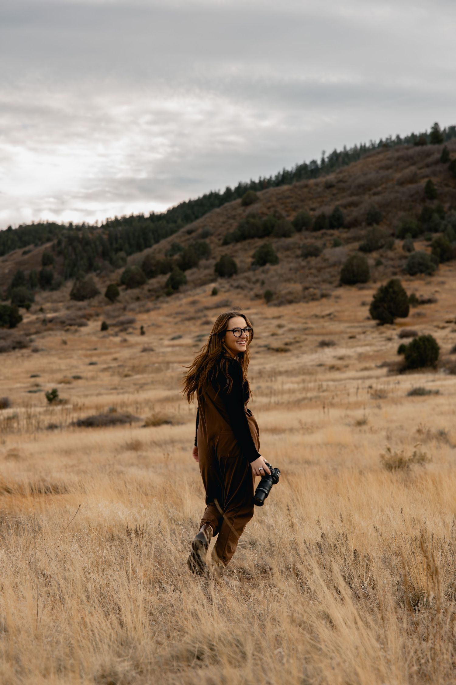Woman in brown vest holding camera smiles in golden field with tree-covered hills under cloudy sky.