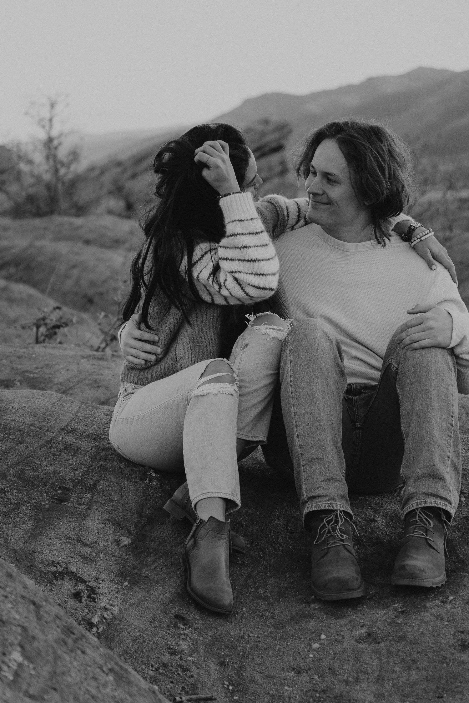 A couple in casual sweaters and jeans sit close together on a hillside against a mountain backdrop in black and white.