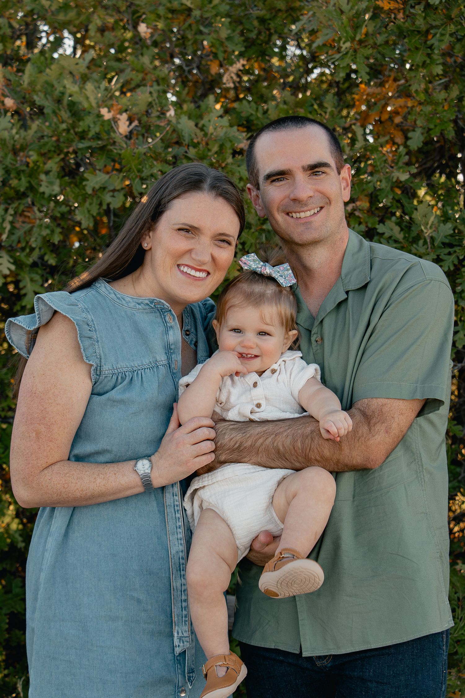 Family in coordinating blue and green outfits poses together outdoors with baby in white dress against leafy background.