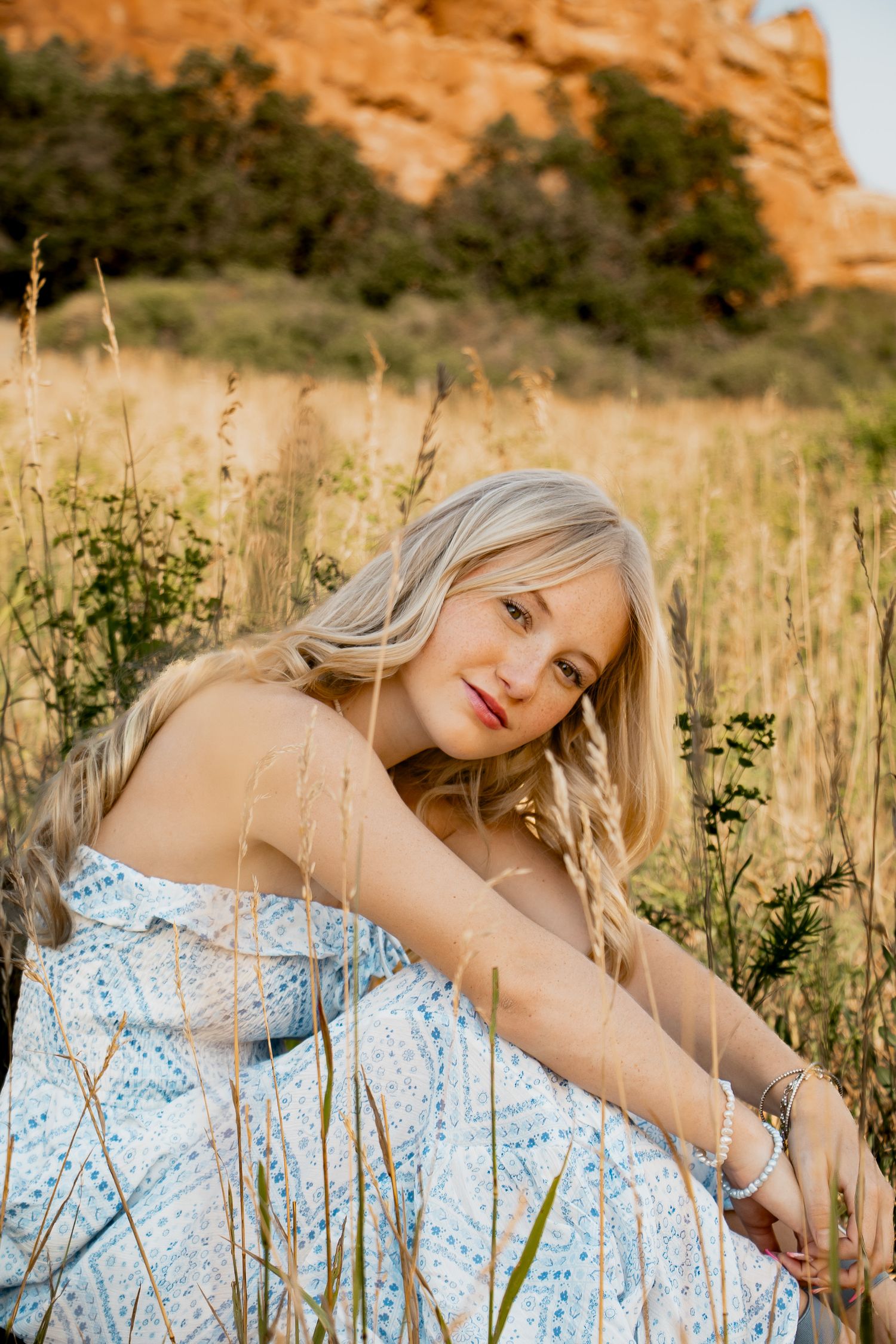A woman in a blue patterned dress poses gracefully in a field with desert red rocks in the background.